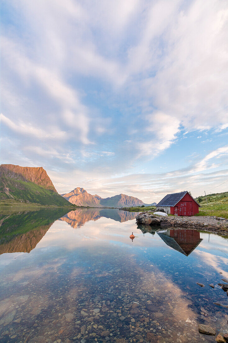 Midnight sun on peaks and typical rorbu reflected in the clear sea at night Vengeren Vagpollen Lofoten Islands Norway Europe