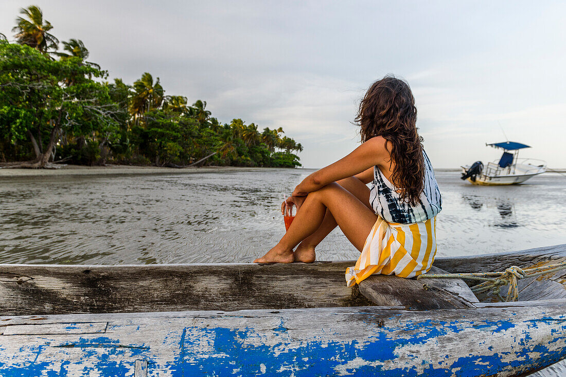 Photograph of young woman sitting in wooden canoe by tropical beach, Boipeba Island, South Bahia, Brazil