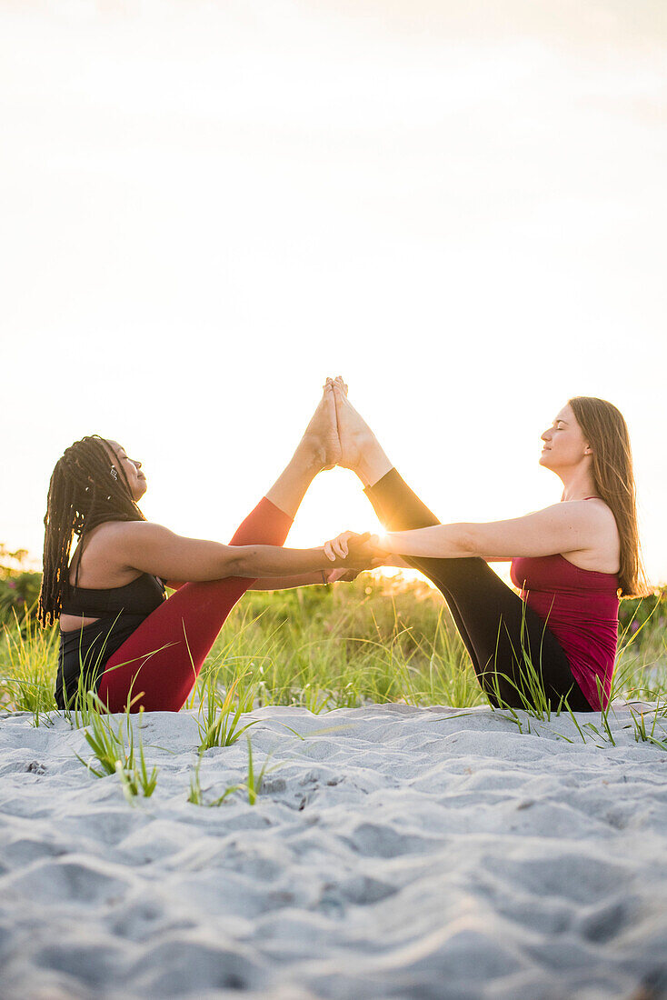 Fotografie von zwei Frauen, die Hände mit den Beinen in einer Luft halten, beim Handeln von Yoga in der unterstützten Boots-Haltung (Navasana), Newport, Rhode Island, USA.