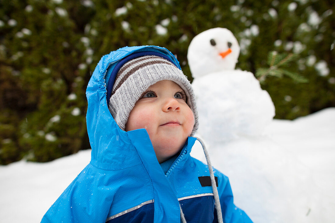 Winter scene with baby boy outdoors with snowman in background, Langley, British Columbia, Canada