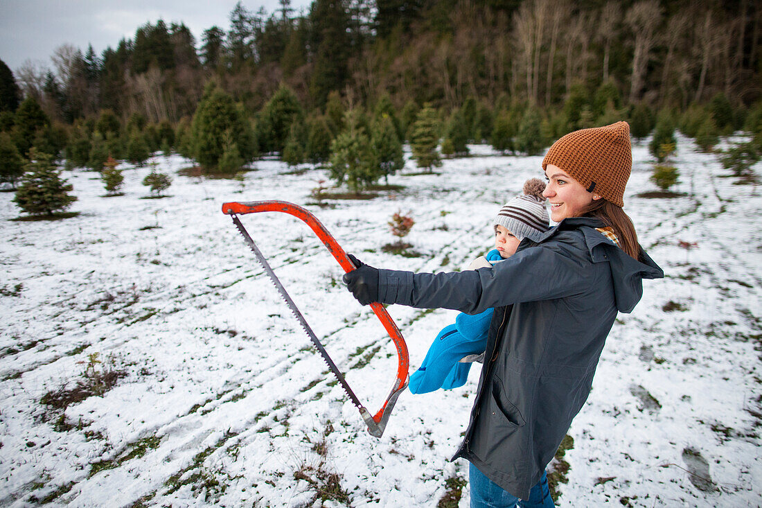 Mutter und Baby, die eine Säge halten und einen Weihnachtsbaum am Bauernhof, Langley, Britisch-Columbia, Kanada vorwählen