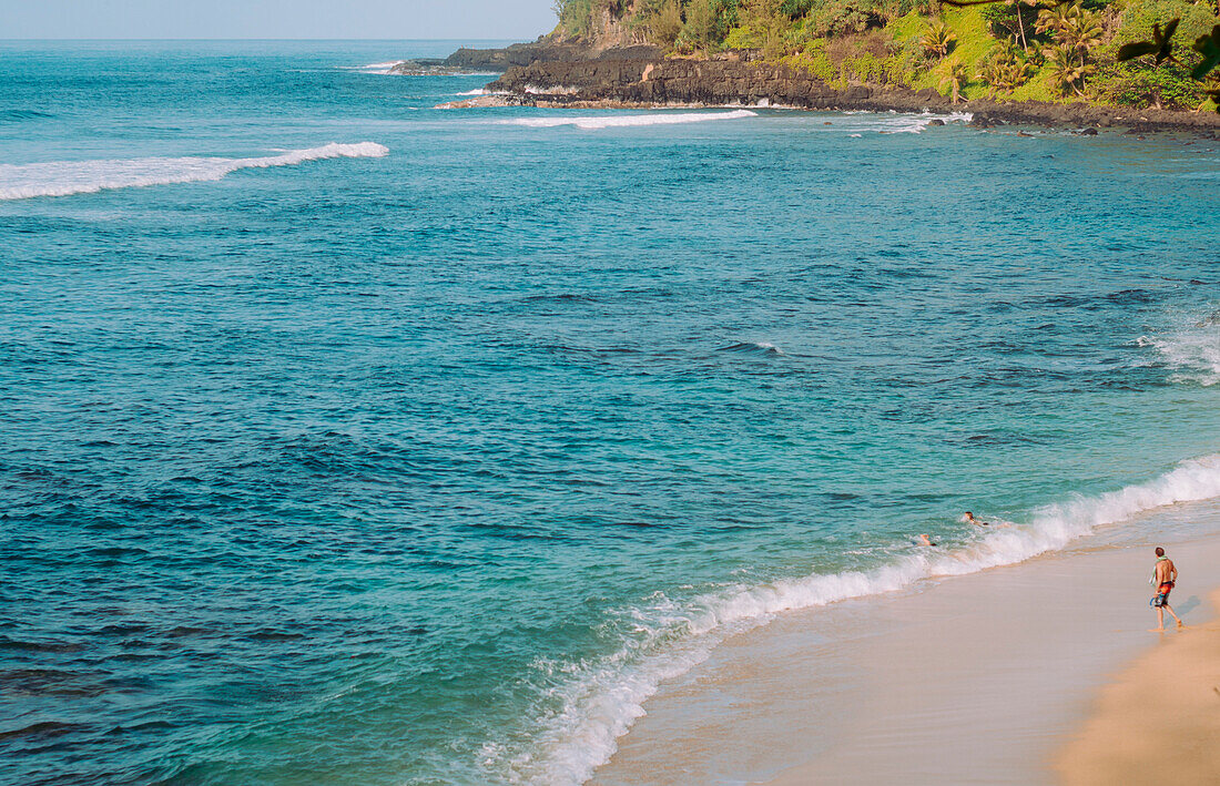 Ein Mann geht an einem Strand in der Nähe von Princeville HI, während zwei Frauen in den Wellen schwimmen