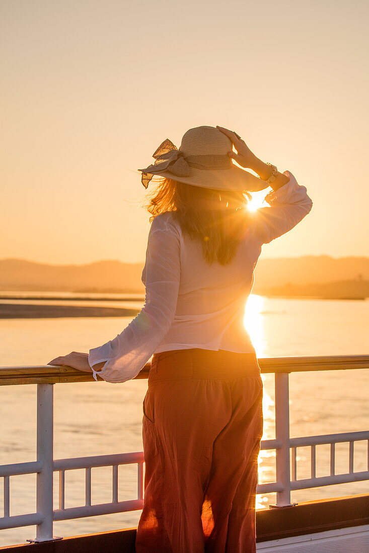 Young blonde woman with sun hat stands at railing of Ayeyarwady (Irrawaddy) river cruise ship Anawrahta (Heritage Line) at sunset, near Bhamo, Kachin, Myanmar