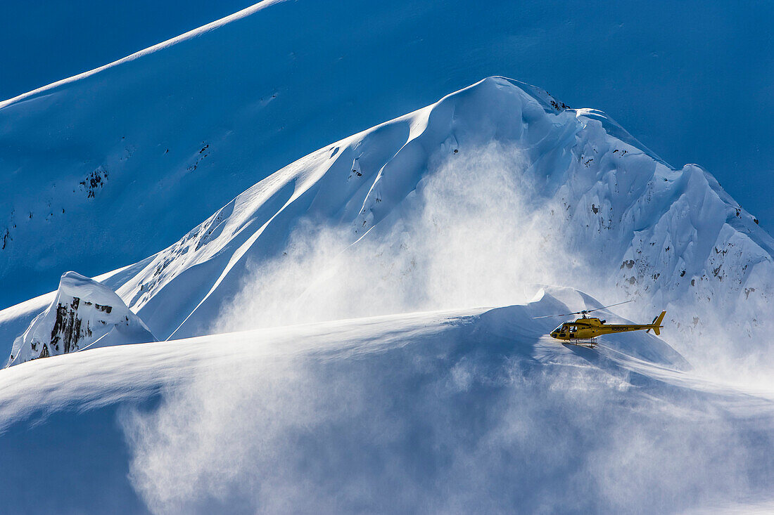 Ein Hubschrauber, der benutzt wird, um Skifahrer und Snowboarder zur Spitze von Bergen zu tragen, hebt ab und bläst Schnee in die Luft an einem sonnigen Drosseltag in Haines, Alaska.