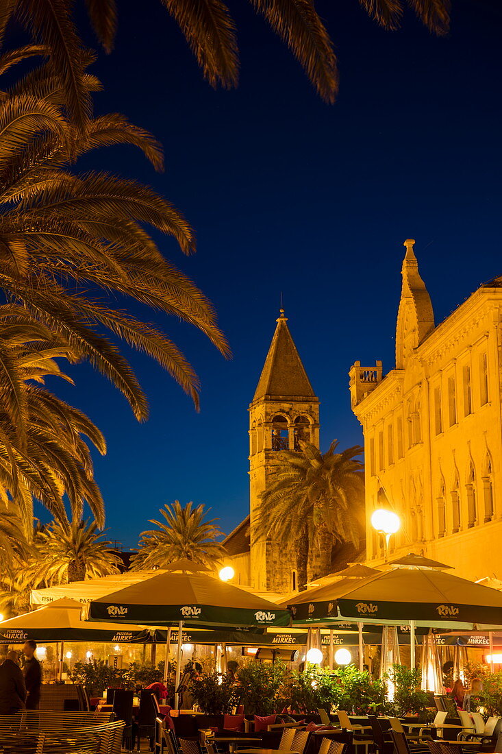 Menschen sitzen vor Restaurants im Freien mit Kirchturm in der Altstadt in der Abenddämmerung, Trogir, Split-Dalmatien, Kroatien, Europa