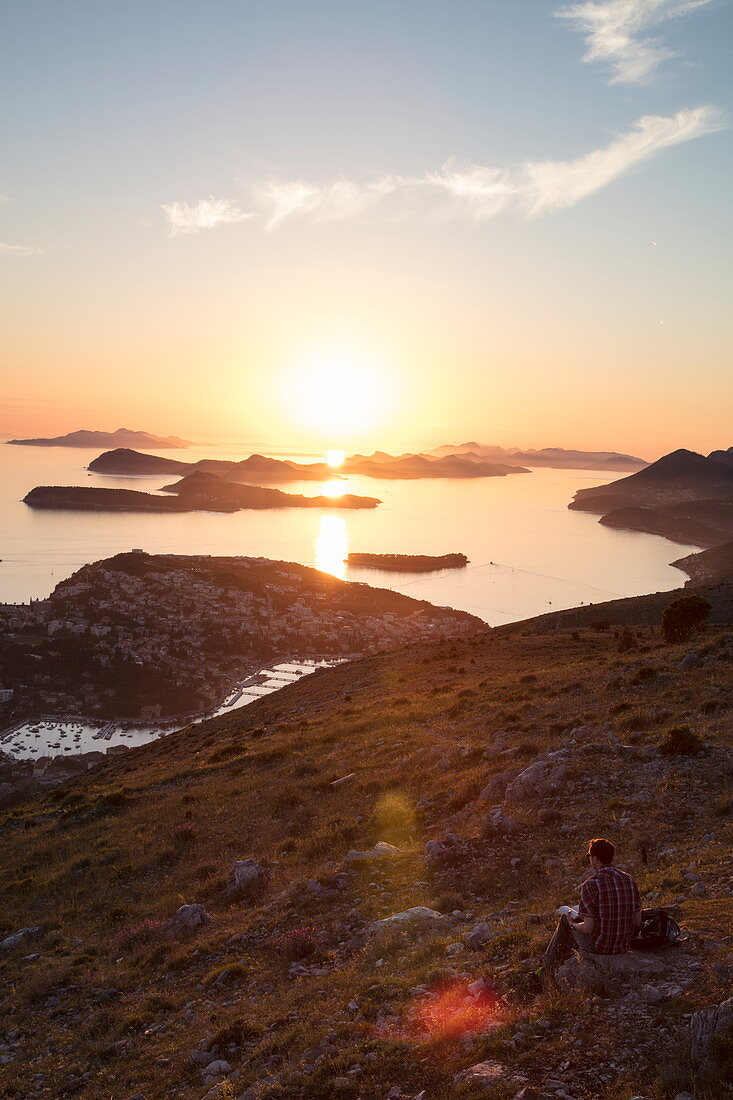 Mann liest Buch mit Blick über die Lapad-Halbinsel und vorgelagerte Inseln, vom Hügel nahe der Bergstatation der Dubrovnik-Gondel bei Sonnenuntergang aus gesehen, Dubrovnik, Dubrovnik-Neretva, Kroatien, Europa