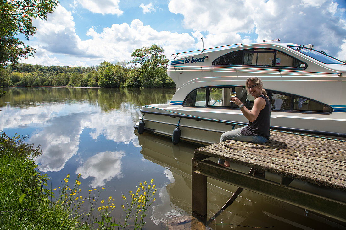 Junge Frau sitz auf Steg und hält Glas mit Weißwein vor Le Boat Elegance Hausboot an Anlegestelle während Bootstörn auf Fluss Petit Saône, Soing, Soing-Cubry-Charentenay, Haute-Saône, Bourgogne Franche-Comté (Burgund), Frankreich, Europa