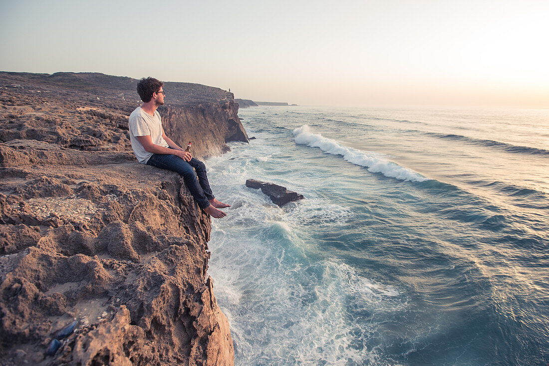 Junger Mann sitzt auf  einem Klippenrand am Meer am Strand Praia da Amoreira,  Aljezur, Faro, Portugal