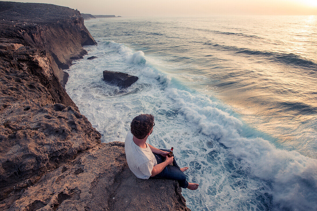 Young man sitting on a cliff edge at the sea at the beach Praia da Amoreira,  Aljezur, Faro, Portugal