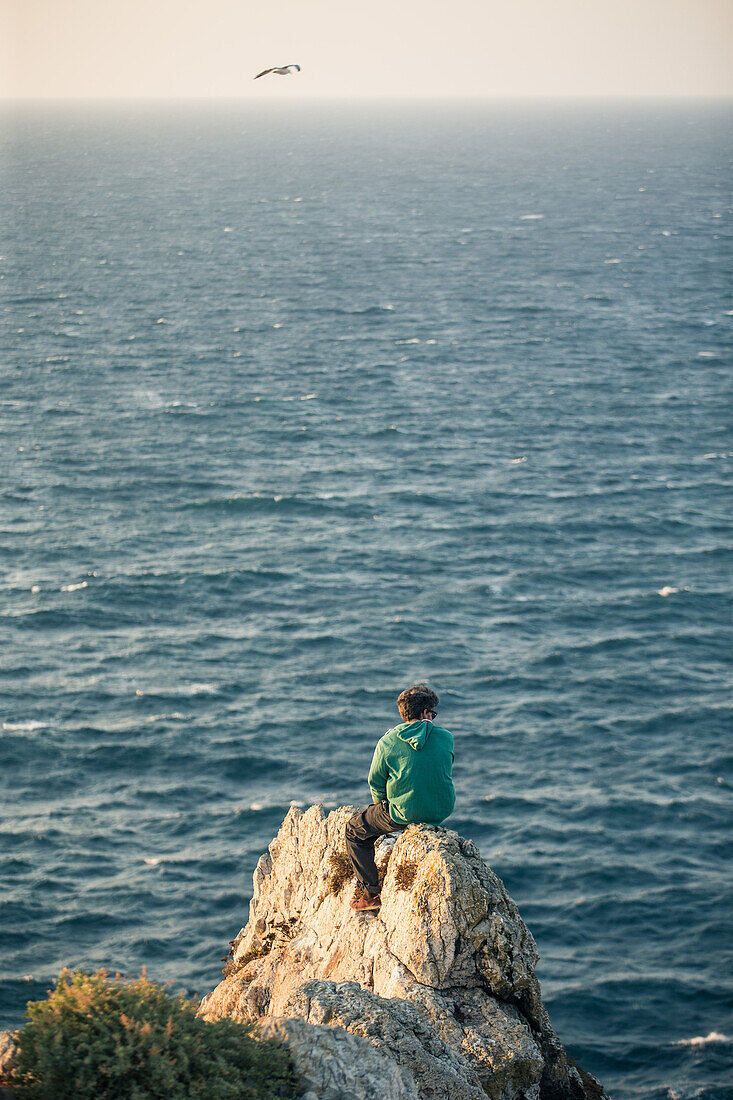 Junger Mann sitzt auf  einem Klippenrand am Meer am Strand Praia da Amoreira,  Aljezur, Faro, Portugal