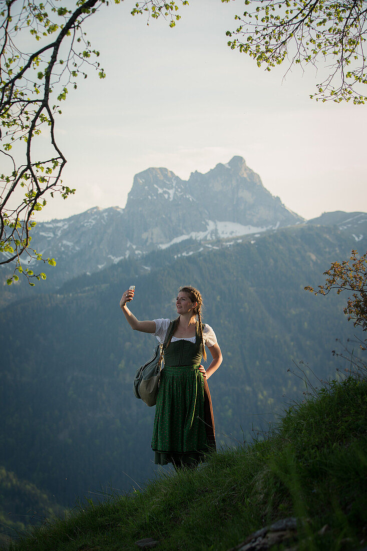 Junge Frau in Tracht macht ein Selfie mit ihrem Handy auf dem Falkenstein im Allgäu, Pfronten, Bayern, Deutschland