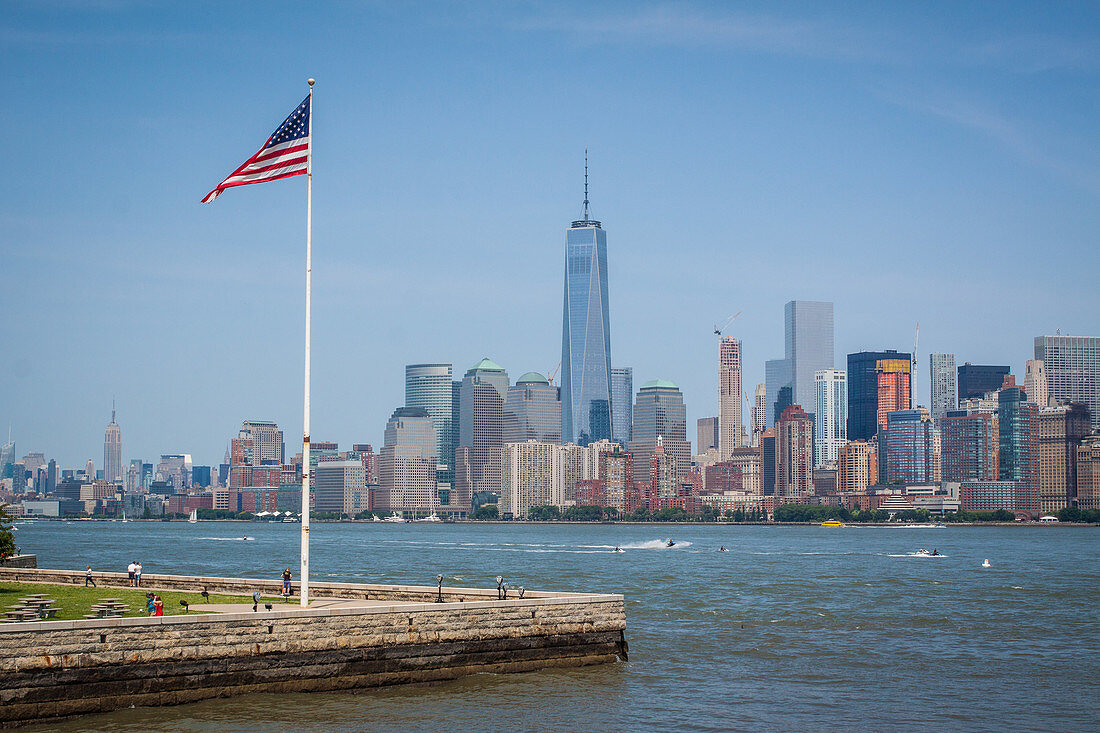 american flag flying over ellis island across from the manhattan skyline with one world trade center and the empire state building in the background, new york harbor, new york city, state of new york, united states, usa