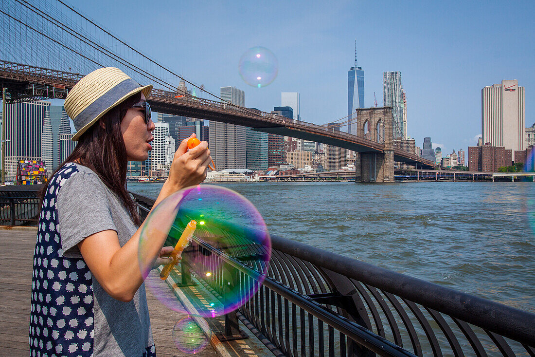 young girl blowing soap bubbles on the waterfront of the east river with the brooklyn bridge and one world trade center in the background, brooklyn bridge park, brooklyn, new york city, state of new york, united states, usa