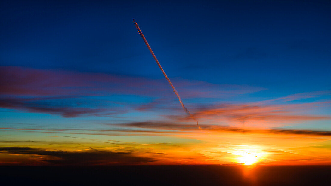 Der Kondensstreifen eines höher fliegenden Flugzeugs wird orange angestrahlt, Sonnenuntergang aus dem Cockpit über Frankreich