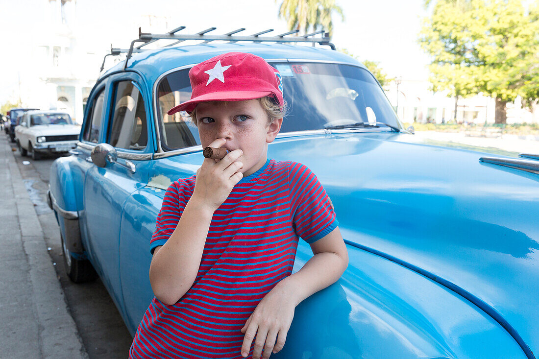 boy standing in front of a blue oldtimer, pretending to smoke a cigarette, at park Parque Jose Marti in the center of Cienfuegos, colonial town, family travel to Cuba, parental leave, holiday, time-out, adventure, MR, Cienfuegos, Cuba, Caribbean island