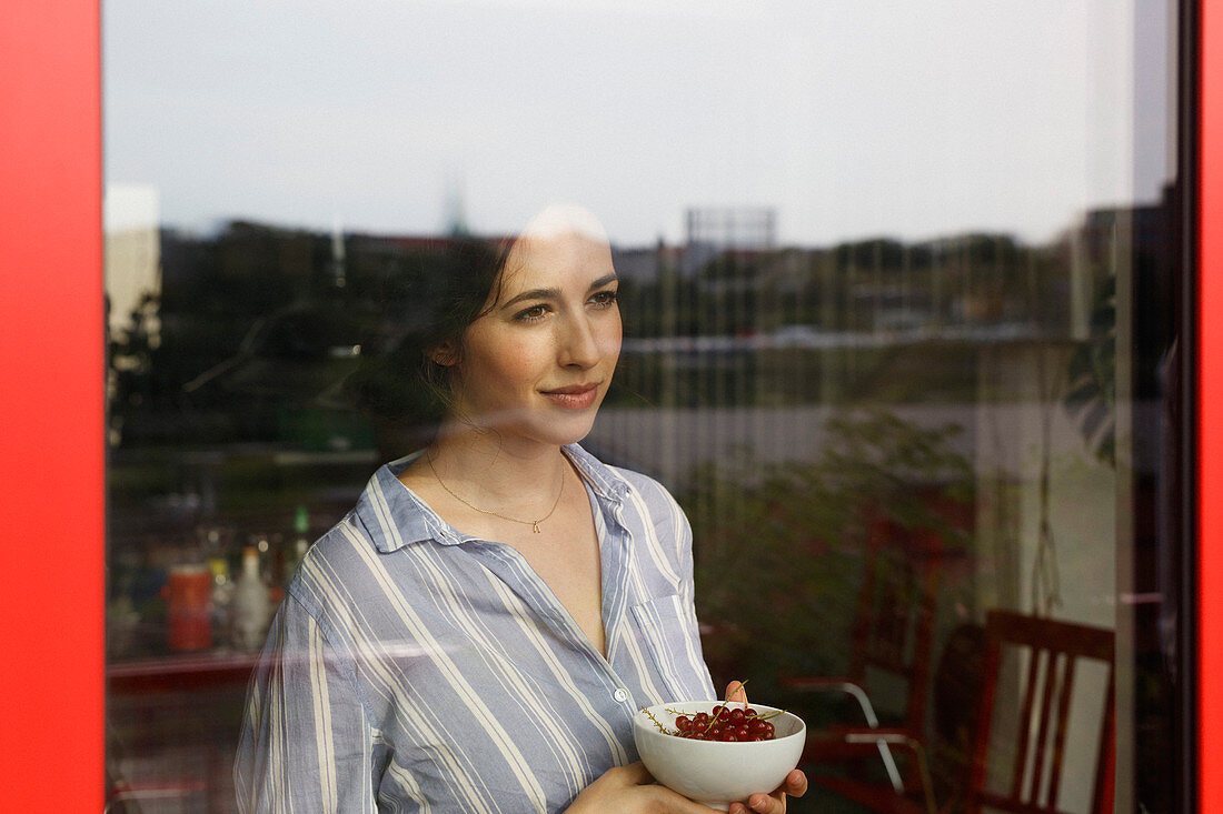 Thoughtful young woman holding bowl with red currants while looking through window