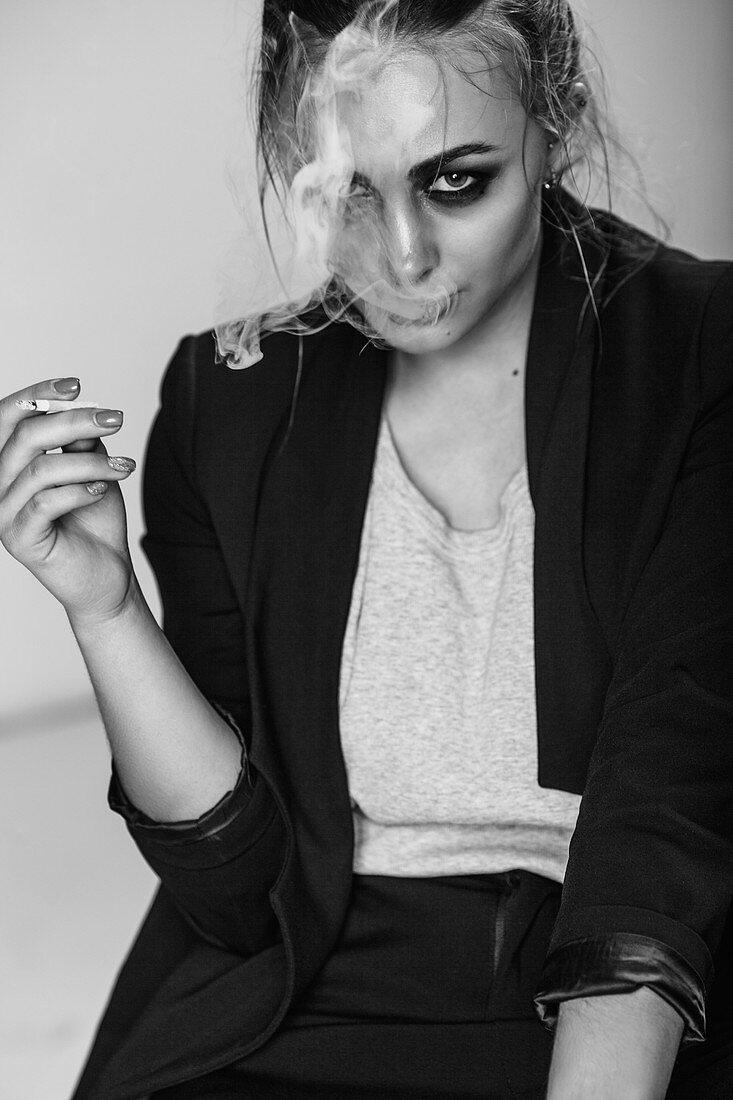 Porträt einer jungen Frau, die im Sitzen eine Zigarette raucht, vor grauem Hintergrund
