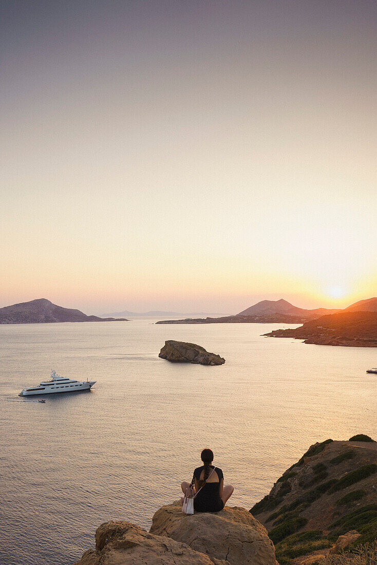 Rear view of woman sitting on rock formation while looking at sea against sky during sunset