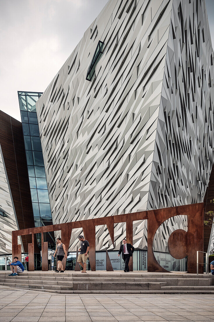 Mann im Anzug läuft durch rostigen Schriftzug TITANIC, moderne Architektur des Schifffahrtsmuseum Titanic City, Belfast, Nordirland, Vereinigtes Königreich Großbritannien, UK, Europa