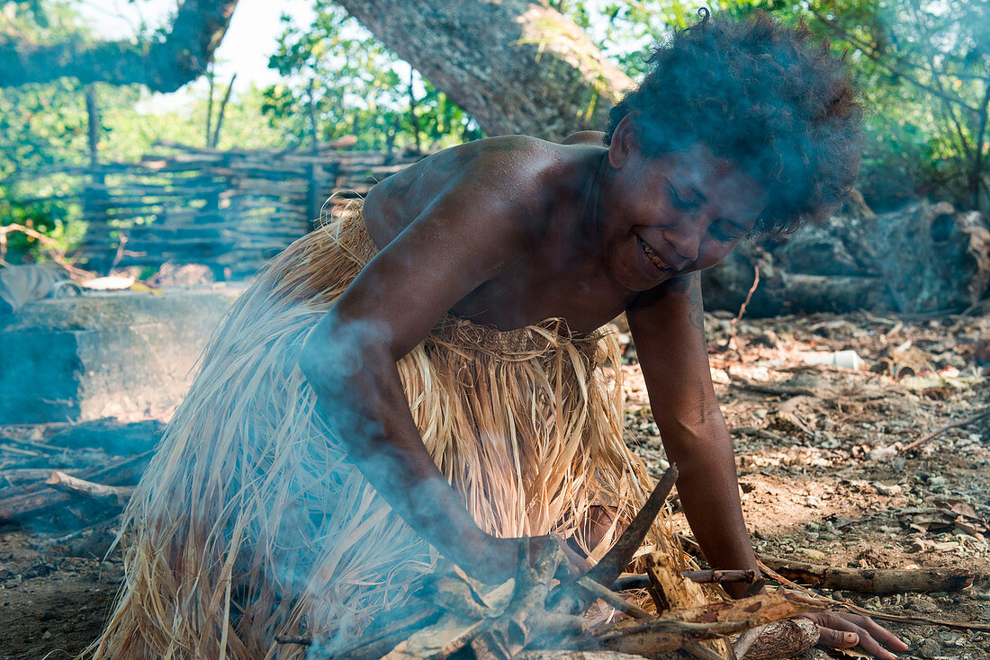 A kneeling woman wearing a traditional grass dress tends her fire during a cultural performance, Arabala, Langa Langa Lagoon, Malaita, Solomon Islands, South Pacific