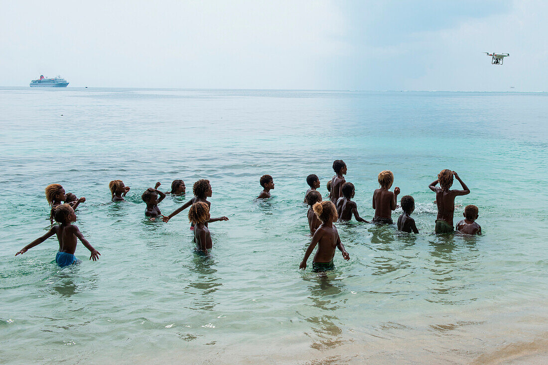 Eine Gruppe Kinder und Jugendliche spielen im Wasser und schauen hoch um eine DJI Phantom 4 Drohne zu beobachten, Santa Ana Island, Makira-Ulawa, Salomonen, Südpazifik