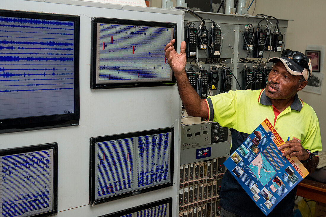 A man in a yellow shirt and hat stands in front of seismic monitoring screens and explains what they measure, Rabaul, East New Britain, Papua New Guinea, South Pacific