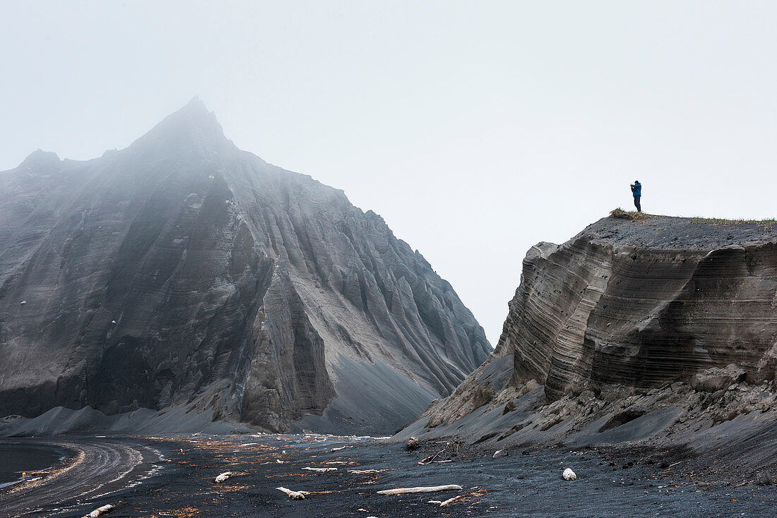 Ein deutscher Tourist fotografiert von einem erodierten Hügel eines schroffen, nebligen Berges nahe der Küste, Atlasova-Insel, Kuril-Inseln, Russland, Asien