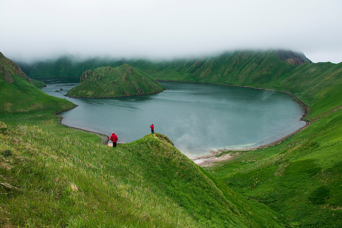 Tourists in red parkas stand on a promontory before a large water-filled caldera, Yankicha Island, Uschischir, Kuril Islands, Sea of Okhotsk, Russia, Asia