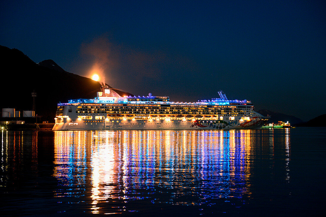 Vollmond steigt über die Berge hinter dem beleuchteten Kreuzfahrtschiff Norwegian Pearl (NCL Cruises) bei der Ausfahrt aus dem Hafen bei Nacht, Juneau, Alaska, USA, Nordamerika