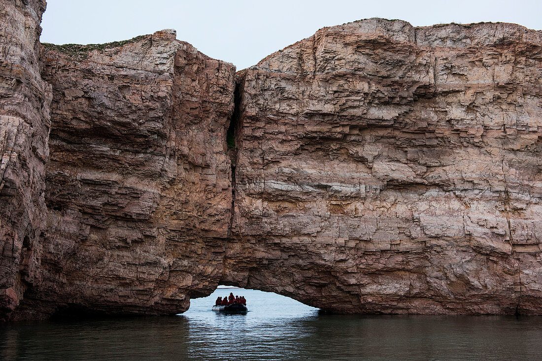 Passengers of expedition cruise ship MS Bremen (Hapag-Lloyd Cruises) in a Zodiac raft pass through a low archway below a large rock with a perpendicular fault in the middle, Pearce Point, Darnley Bay, Northwest Territories, Canada, North America