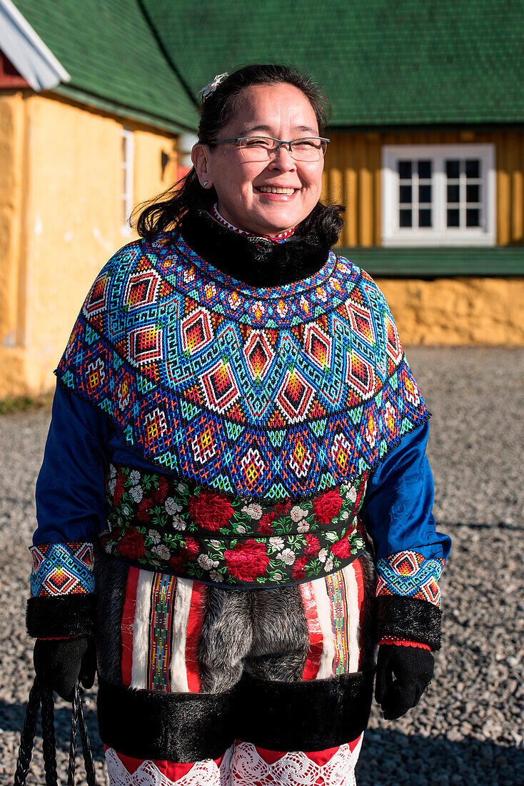 A woman in traditional dress smiles in front of a ocher-colored building with a green roof, Sisimiut, Qeqqata, Greenland