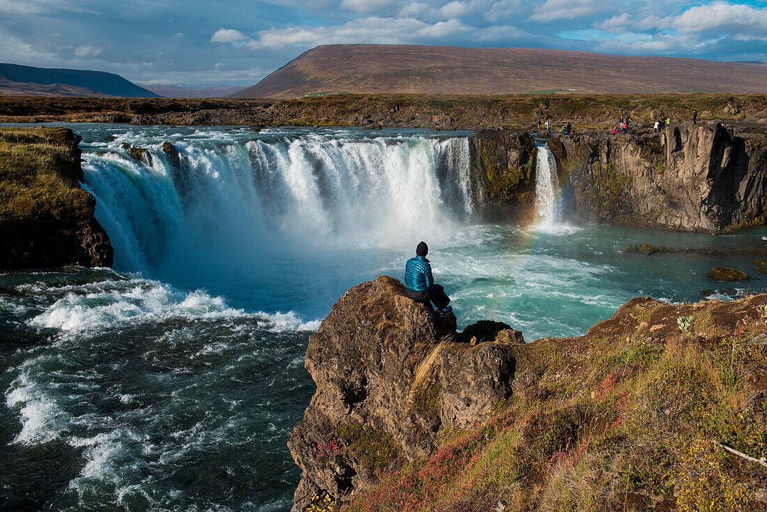 Ein Tourist sitzt auf einem Felsvorsprung und blickt auf den 12 Meter hohen Goðafoss (Wasserfall der Götter), in der nahe Seyðisfjörðdur, im Osten von Island, Europa