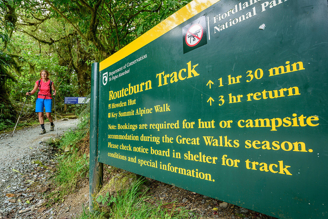 Wanderwegweiser für Routeburn Track, Frau beim Wandern im Hintergrund, Routeburn Track, Great Walks, Fiordlands Nationalpark, UNESCO Welterbe Te Wahipounamu, Queenstown-Lake District, Otago, Südinsel, Neuseeland