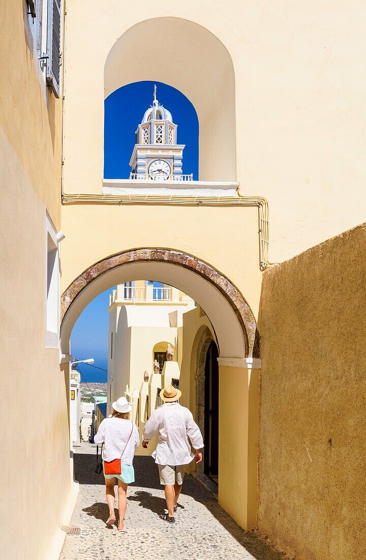 A couple walk under an archway near the St. John the Baptist Cathedral in Fira, Santorini, Cyclades, Greece.