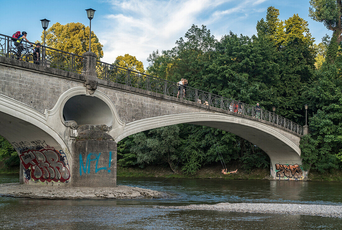 A young man swinging under the Kabelsteg bridge at the Praterinsel at river Isar, passer-by cross the bridge and stay for a while, Munich, Upper Bavaria, Germany