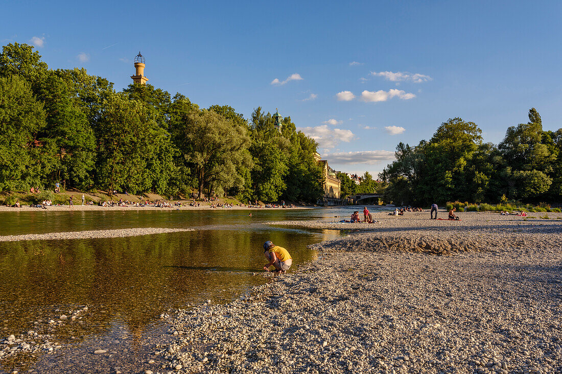 A boy playing in the water on a gravelbank besides the Muffathalle, swimmers and visitors enjoying the evening sun, Muffathalle and Ludwigsbruecke bridge in the background, Munich, Upper Bavaria, Germany