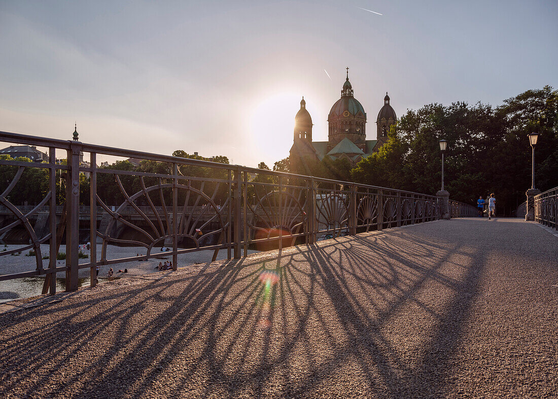 Sonnenuntergang hinter der Lukaskirche, ein Junge rennt über den Kabelsteg, Menschen sitzen an der Isar auf einer Kiesbank, München, Oberbayern, Deutschland