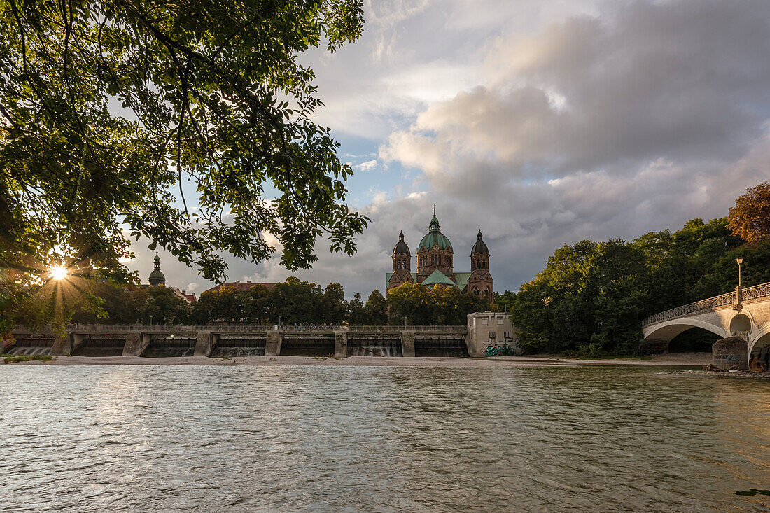 Blick über die Isar auf das Wehr und St. Lukas bei Sonnenuntergang, rechts der Kabelsteg, München, Oberbayern, Deutschland