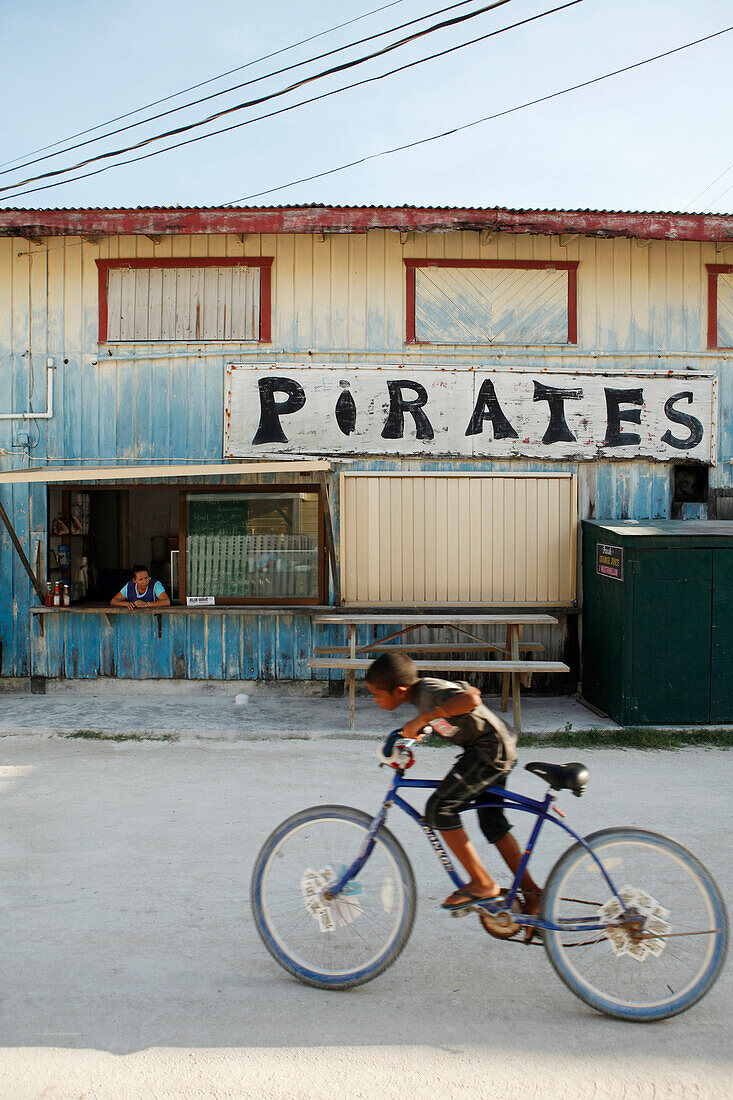 BELIZE, Caye Caulker, a boy rides his bike past pirates restaurant