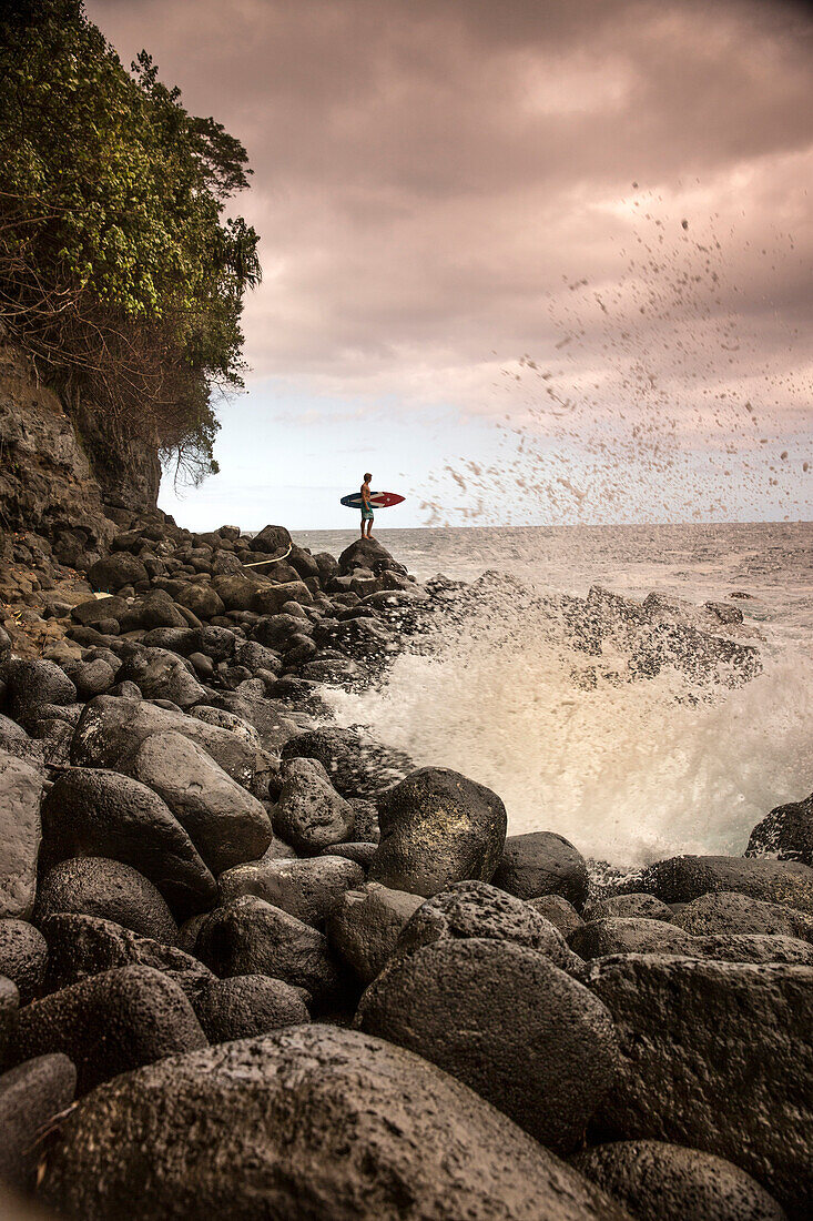 USA, Hawaii, The Big Island, surfer watches the waves on a rocky shoreline at the Hakalau River mouth