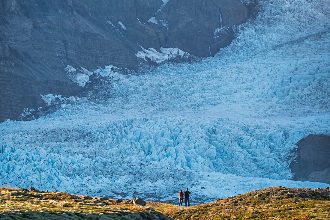 A Couple Standing In Front Of A Glacier Along The South Coast Of Iceland, Admiring Nature At Its Best; Iceland