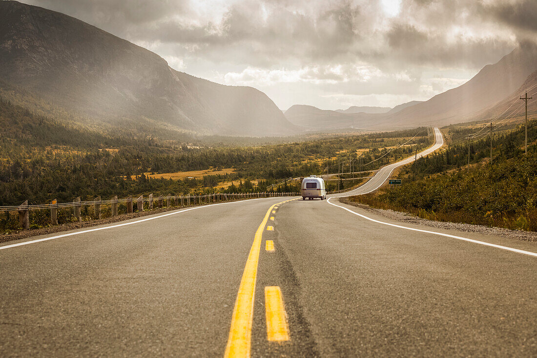 A vehicle with trailer heads down a road leading through fields and mountains under a cloudy sky; Newfoundland, Canada