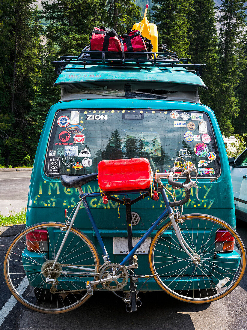 A camper van in a parking lot covered with bumper stickers and graffiti, and with a luggage on a rack and bike on the back; Field, Alberta, Canada