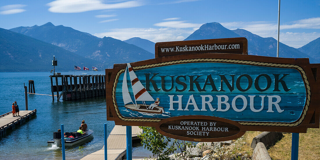 A sign for Kuskanook Harbour on Kootenay Lakewith people on a dock and in a boat in the background; Creston, British Columbia, Canada