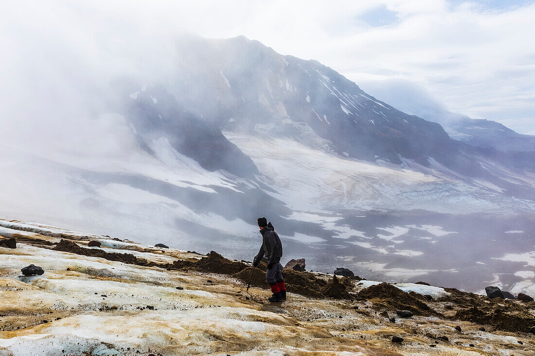 A man looks toward Trident Volcano through clouds while ascending the Knife Creek Glaciers in Katmai National Park; Alaska, United States of America