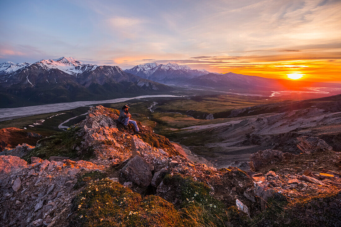 A man observes a tranquil sunset from an alpine perch high above the Delta River in the Alaska Range; Alaska, United States of America