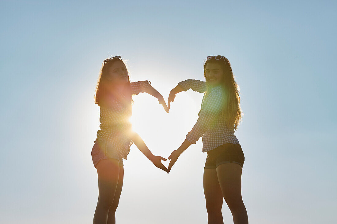 Two teenage girls stand making the shape of a heart with their arms as the sunlight shines through, Woodbine Beach; Toronto, Ontario, Canada