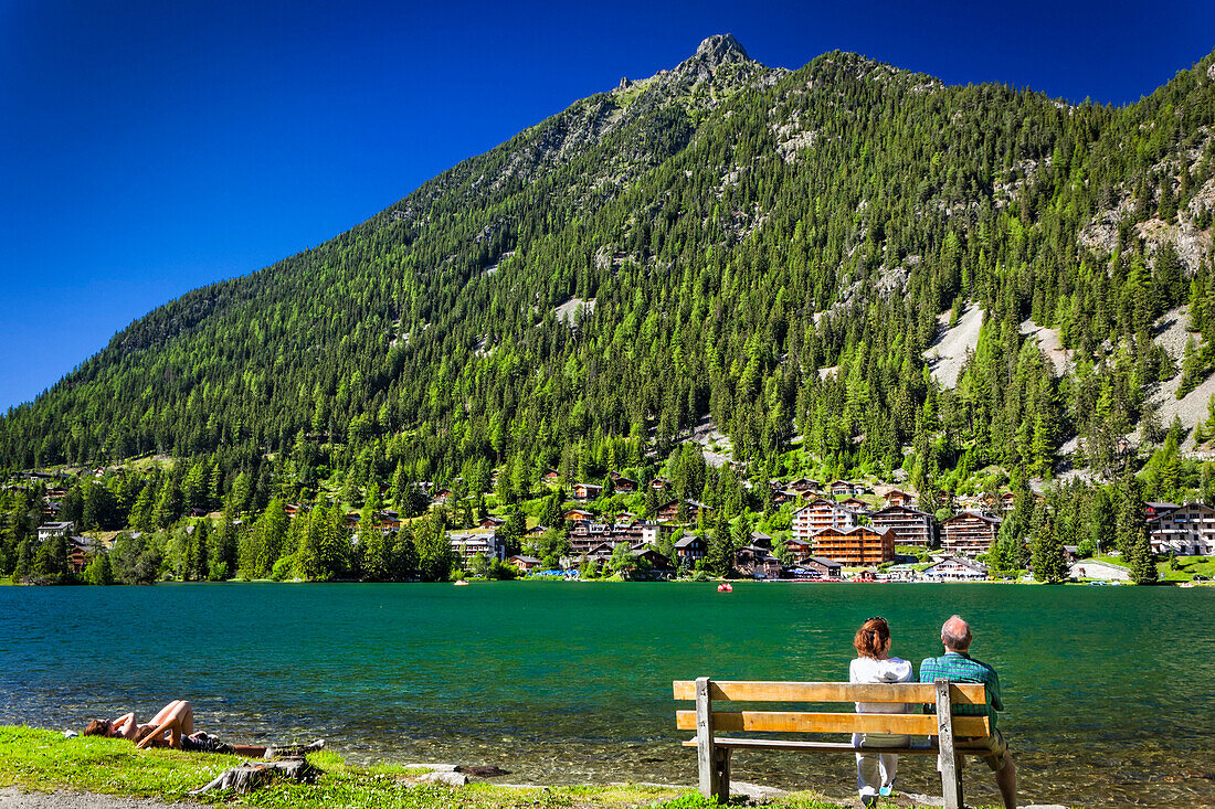 A couple enjoys the view on a bench at Champex Lake shore under blue sky, mountain and resorts are in the background; Champex, Valais, Switzerland