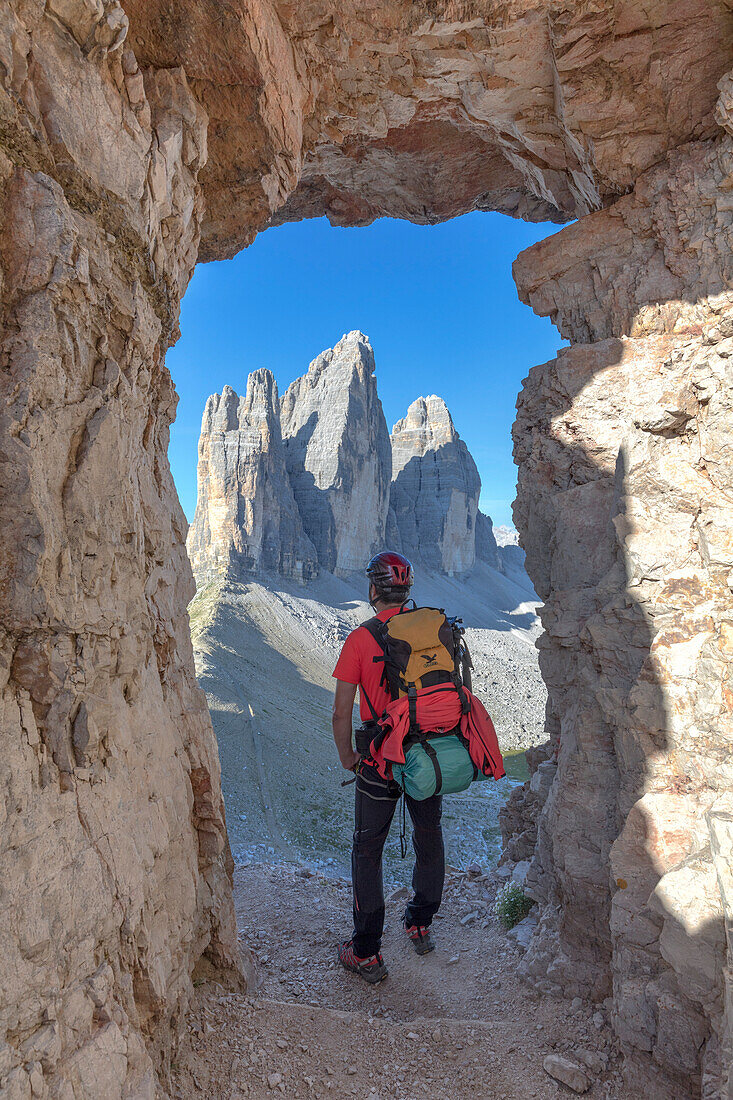 Hiker looking the Tre Cime di Lavaredo / Drei Zinnen through a window in the rock at the foot of the mount Paterno / Paternkofel, Sexten Dolomites, South Tyrol, Bolzano, Italy