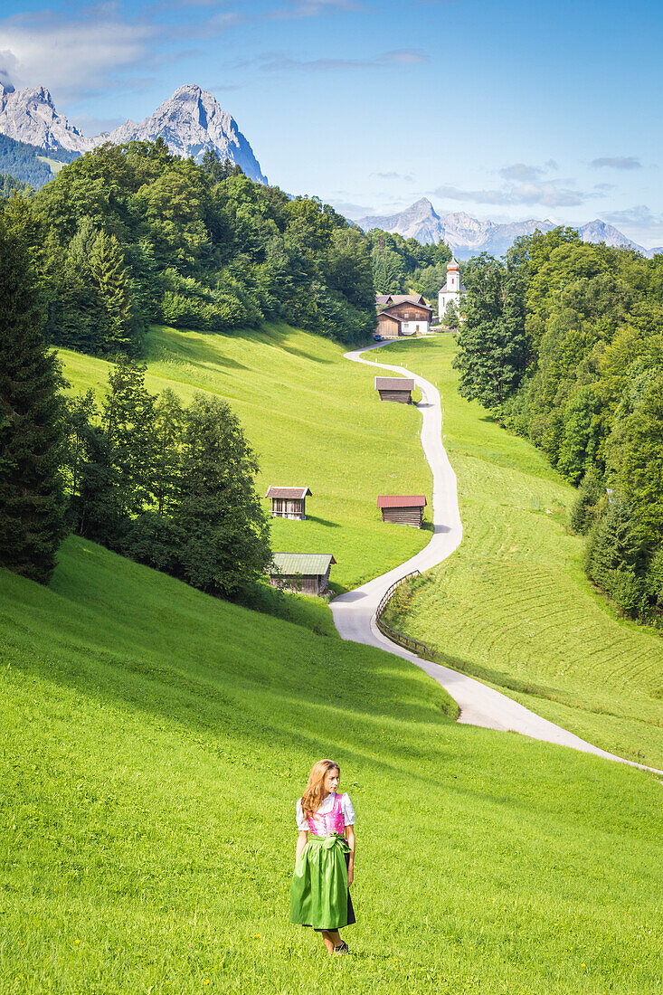 A girl in Typical Bayern dress walking in front of Wamberg village, with Mount Zugspitze and Waxenstein on the background, Garmisch Partenkirchen, Bayern, Germany